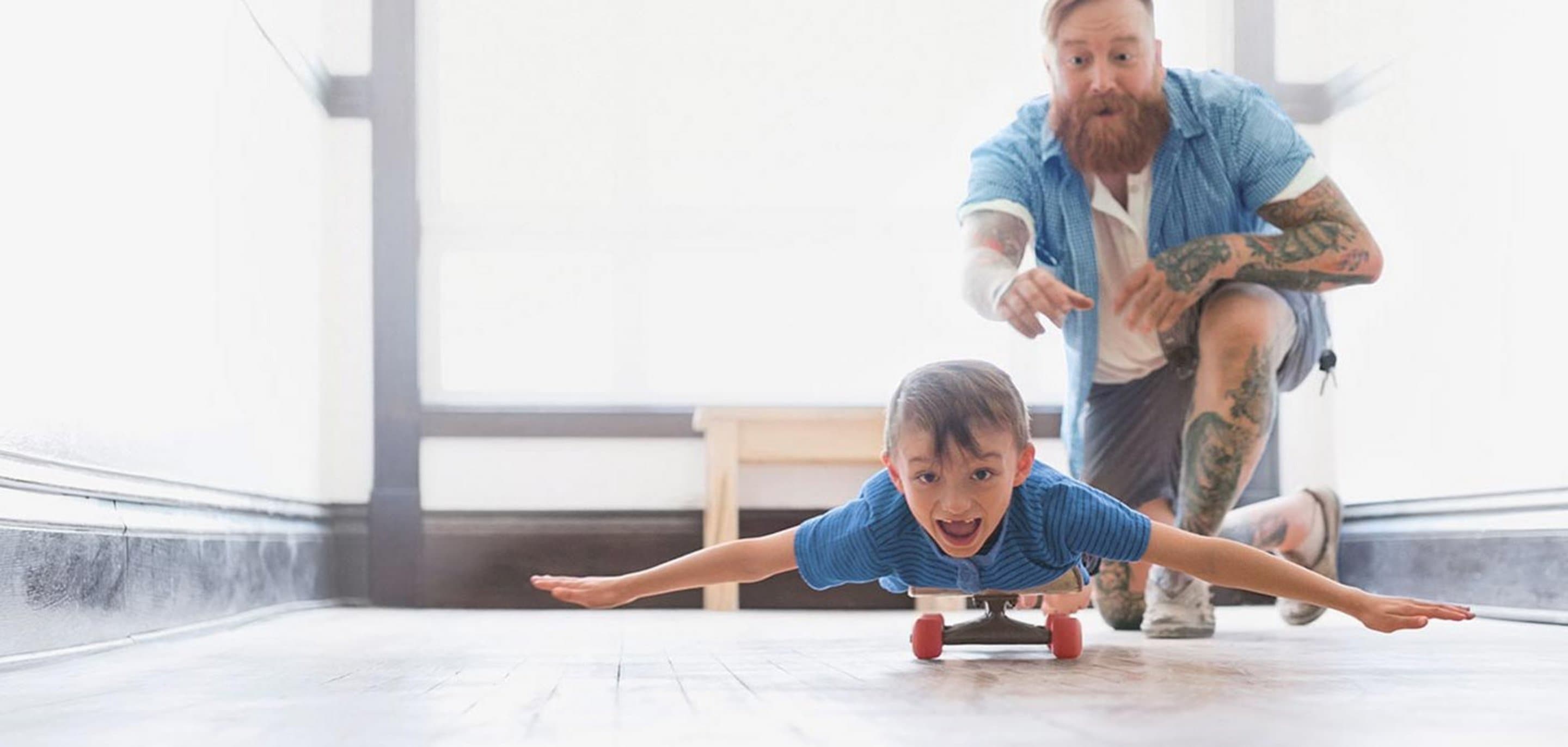 Vater schubst Sohn an, der mit ausgebreiteten Armen auf einem Skateboard liegt.