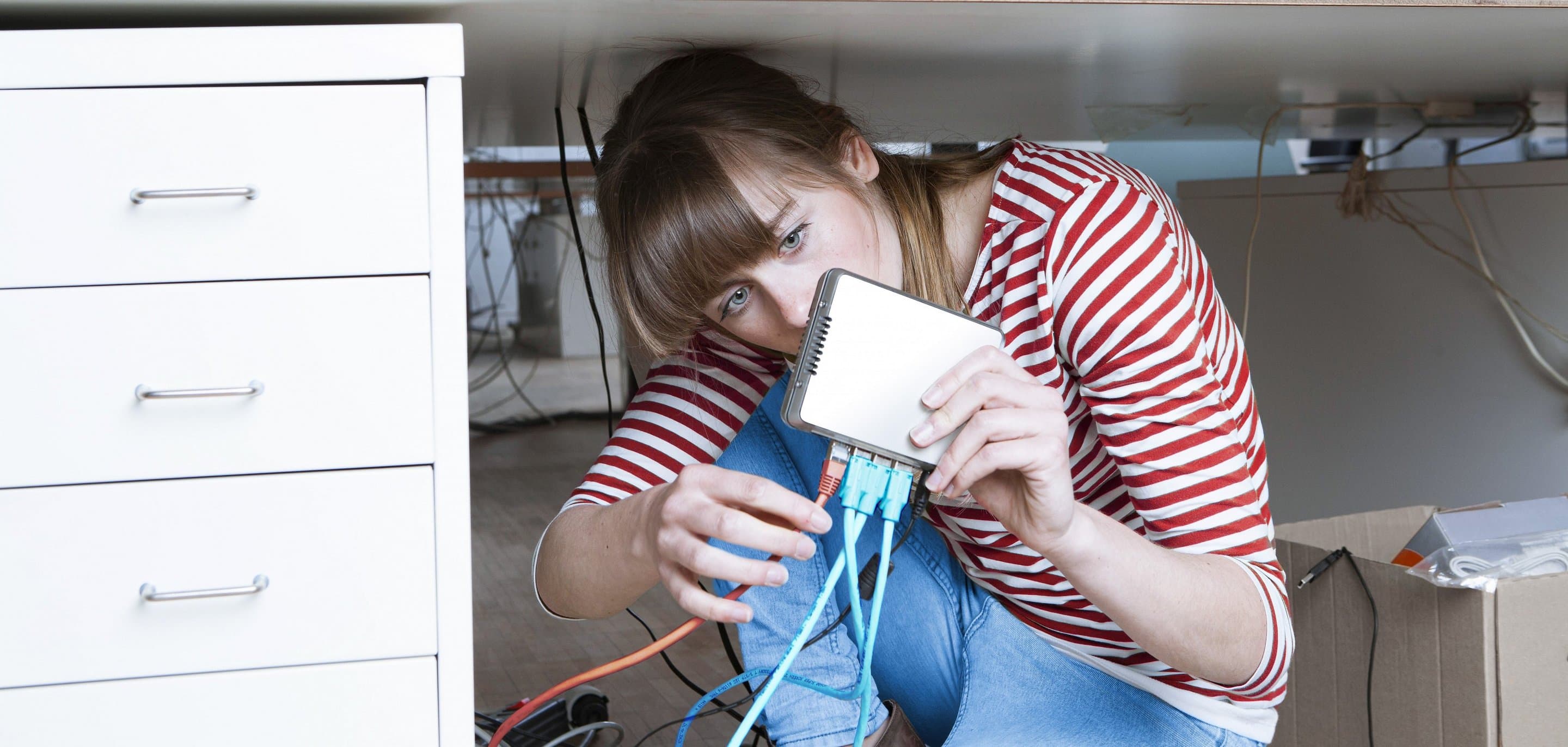 Frau sitzt unter einem Schreibtisch und steckt ein LAN-Kabel in einen Router