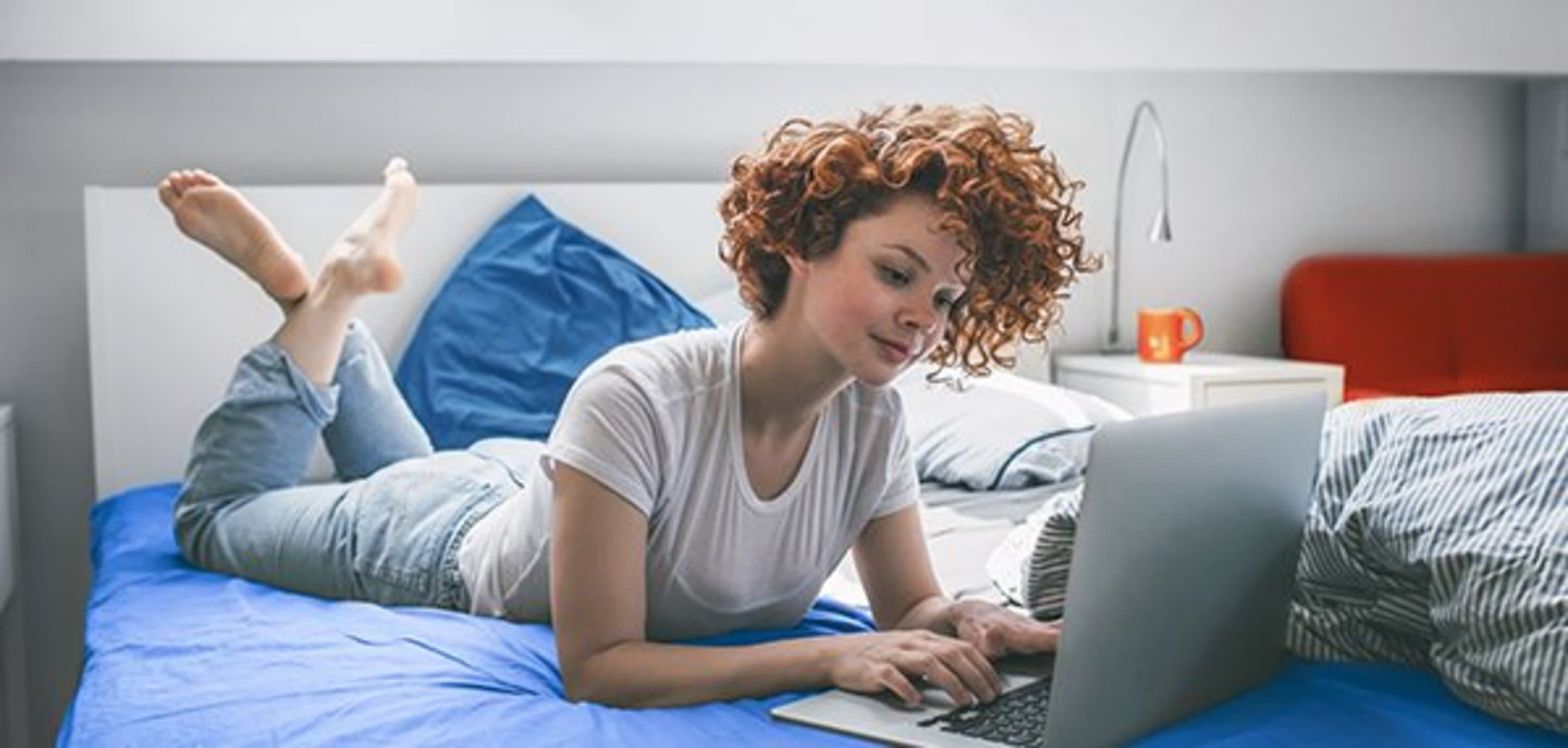 Frau mit Locken liegt auf dem Bauch falsch herum im Bett und surft am Laptop im Internet.