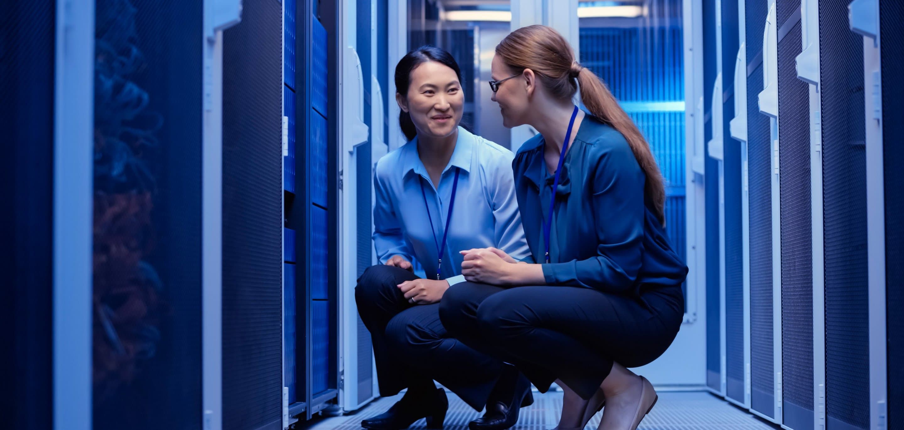 Rechenzentrum Zwei Personen in Businesskleidung knien in einem blau beleuchteten Serverraum und unterhalten sich zwischen Serverracks.