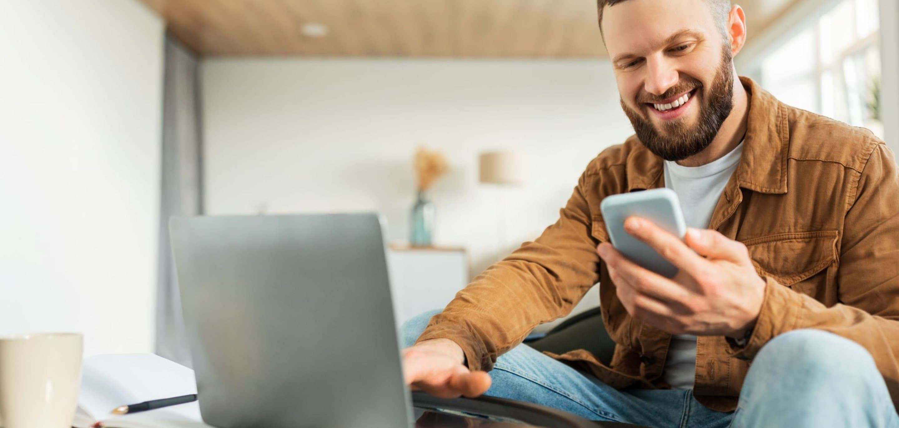 Person sitzt in einem modernen Raum vor einem Laptop und hält ein Smartphone in der Hand, auf dem Tisch steht eine helle Kaffeetasse.