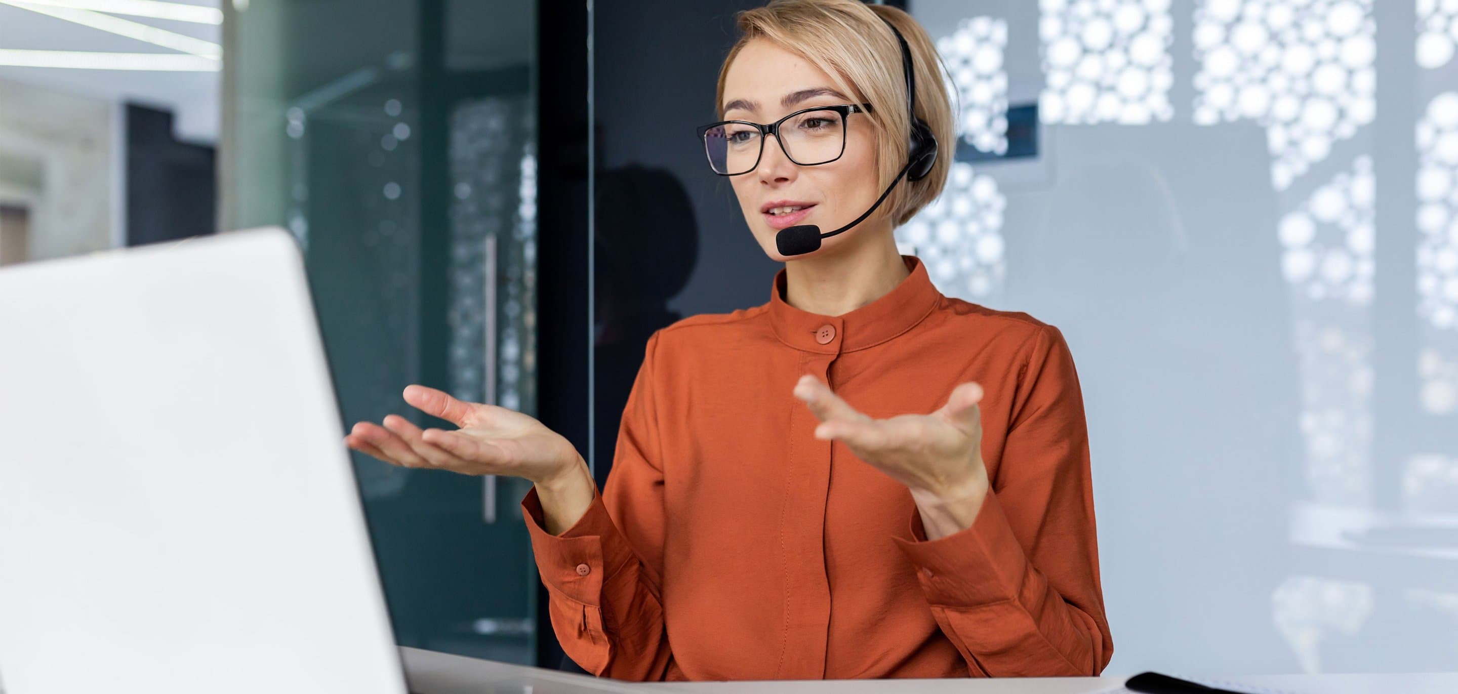 Kundenservice Person mit orangefarbenem Shirt und Headset sitzt am Schreibtisch vor einem Laptop, im Hintergrund Glas- und Musterfenster.