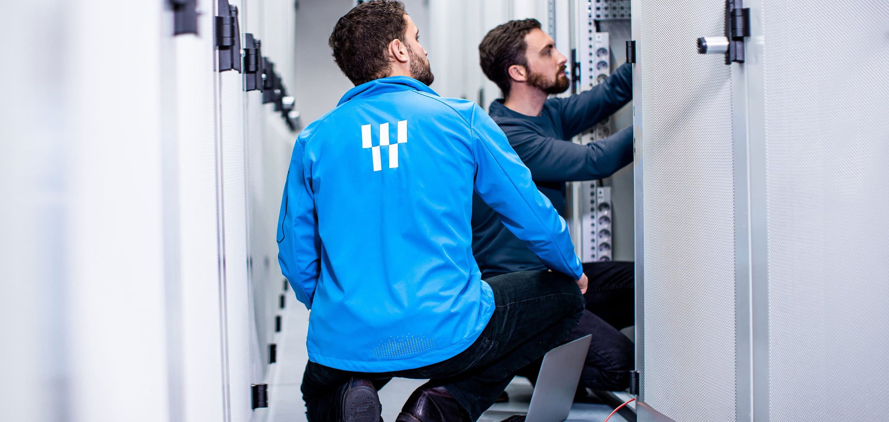 Zwei Personen arbeiten in einem Serverraum an offenen Serverschränken, eine trägt eine blaue Jacke mit NetAachen-Logo, auf dem Boden liegt ein Laptop.