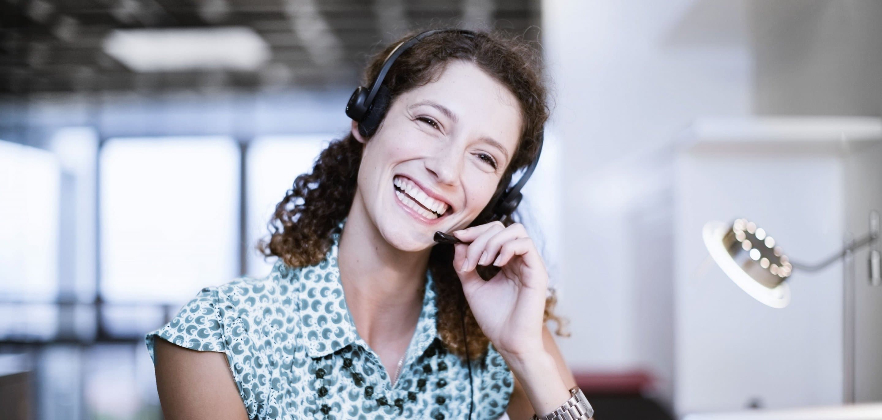 Person trägt ein Headset und sitzt an einem Schreibtisch in einem modernen Büro, im Vordergrund eine Schreibtischlampe.
