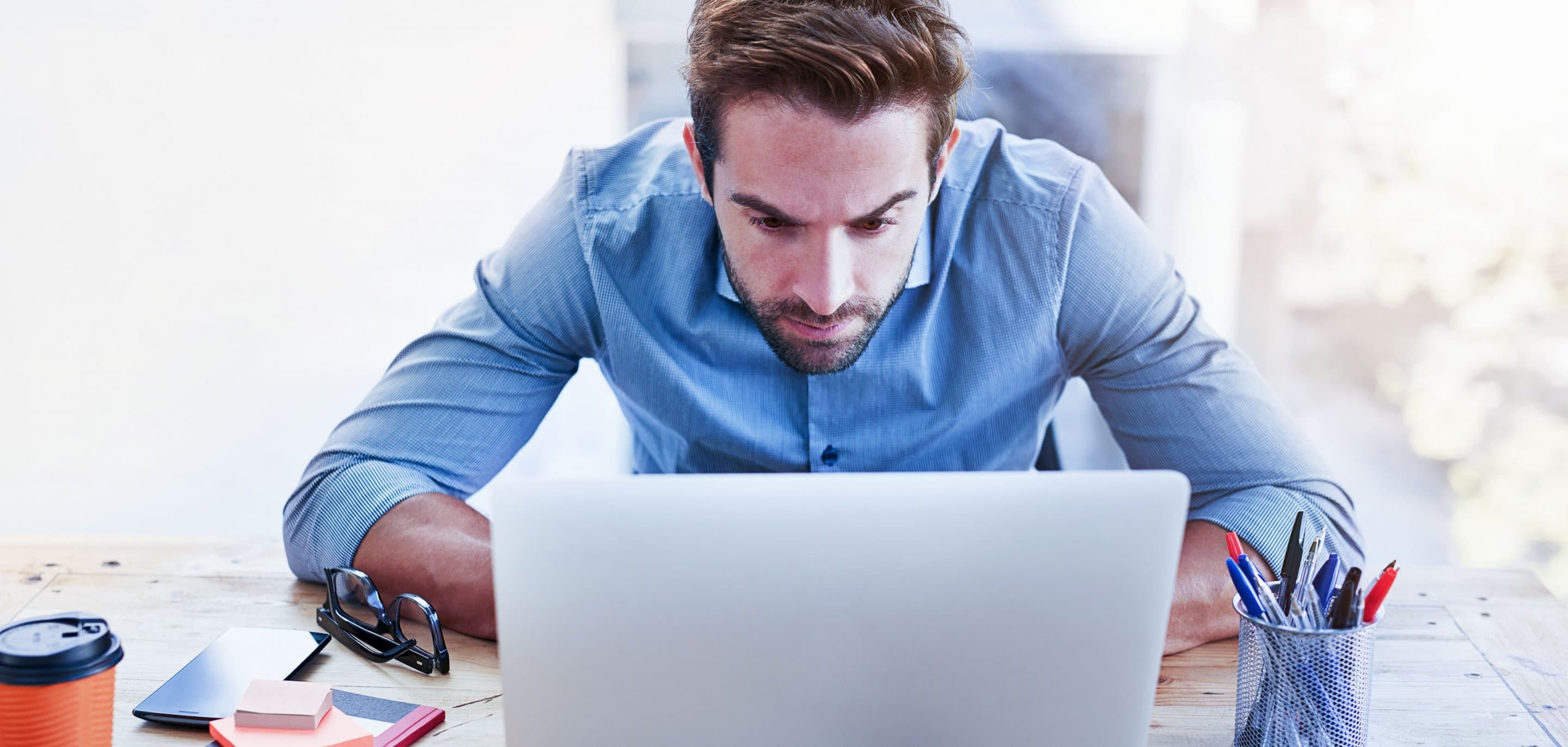 Person in hellblauem Hemd sitzt an einem Schreibtisch vor einem Laptop, daneben liegen eine orangefarbene Kaffeetasse, ein Smartphone und ein Stiftehalter.
