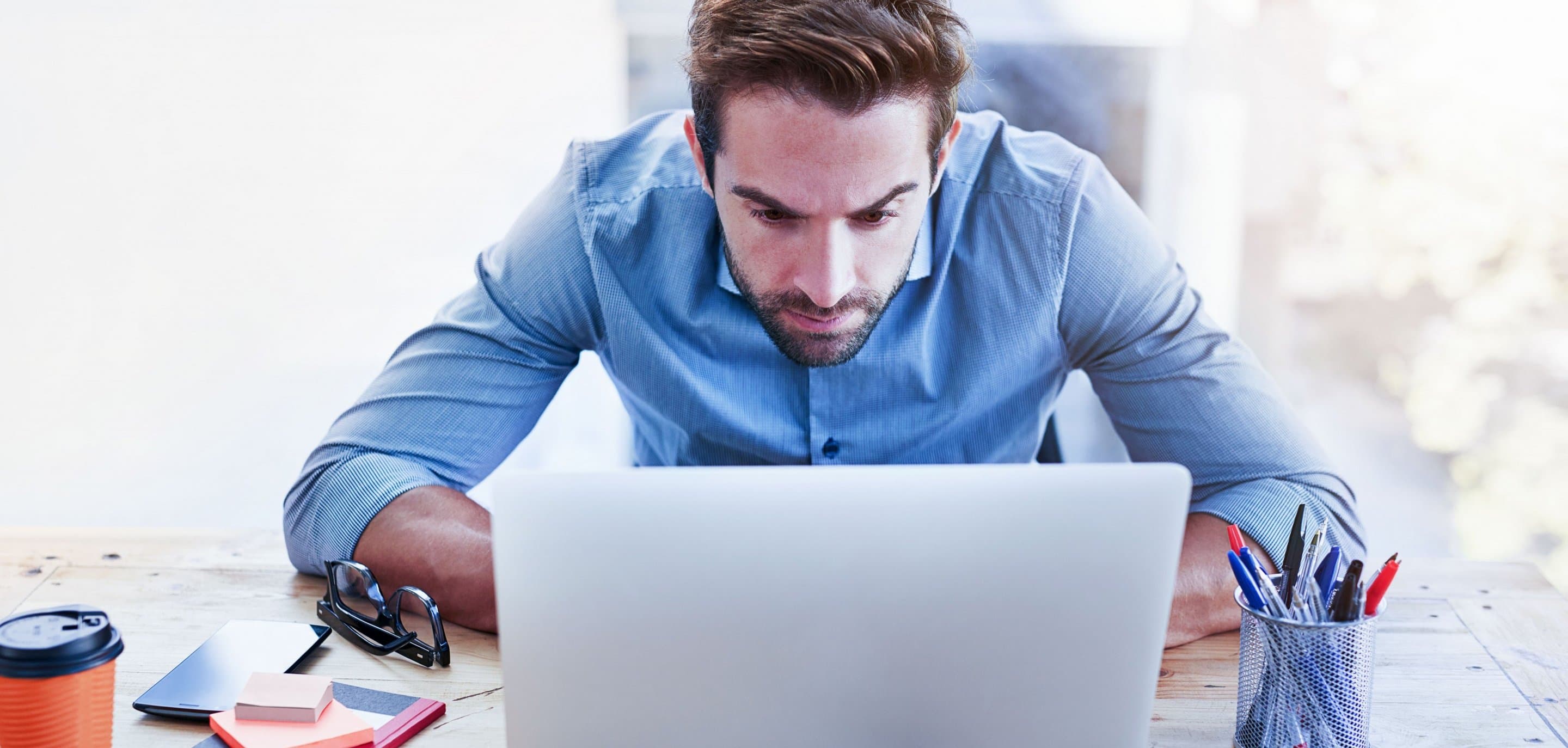 Person in hellblauem Hemd sitzt an einem Schreibtisch vor einem Laptop, daneben liegen eine orangefarbene Kaffeetasse, ein Smartphone und ein Stiftehalter.