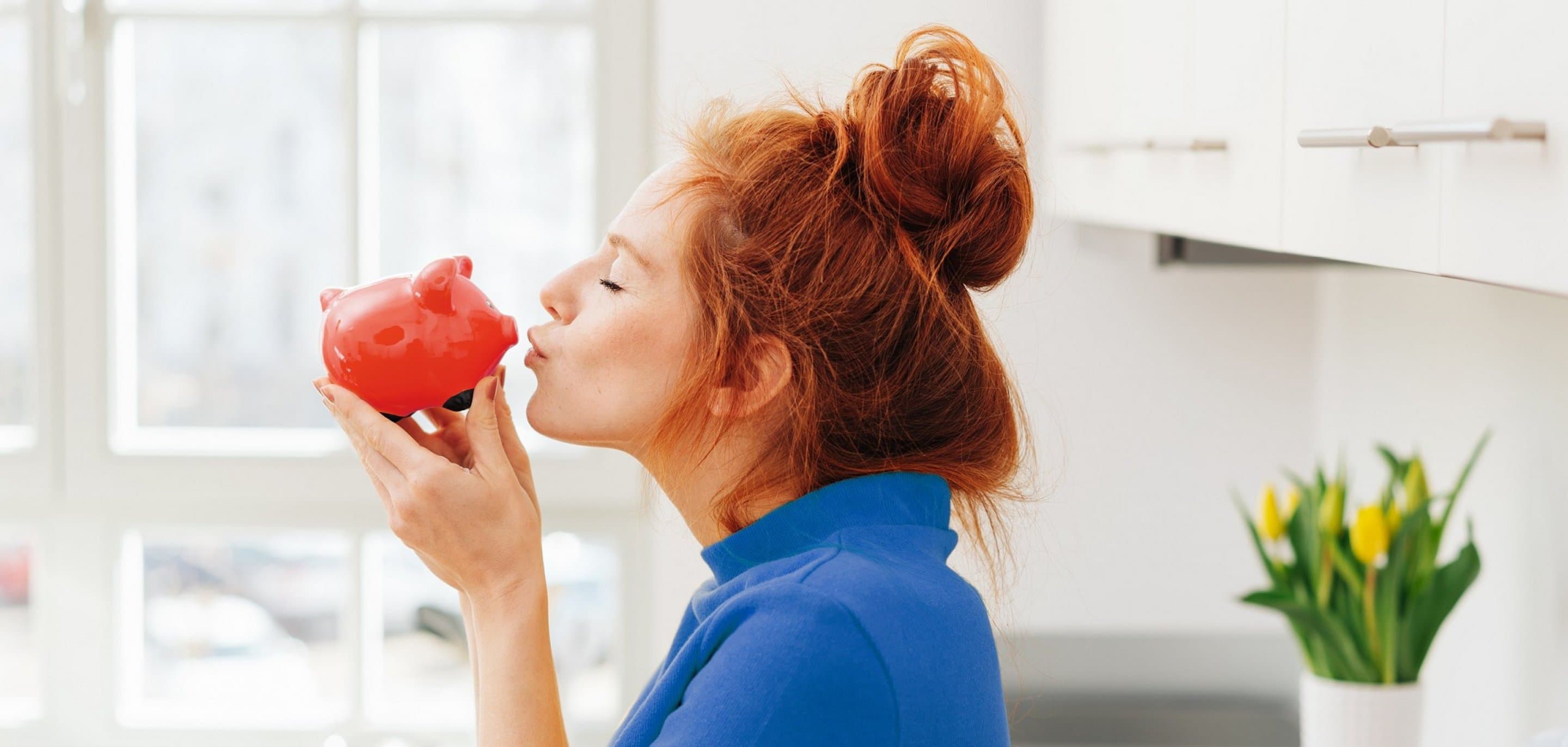Rothaarige Frau mit Dutt küsst rotes Sparschwein.