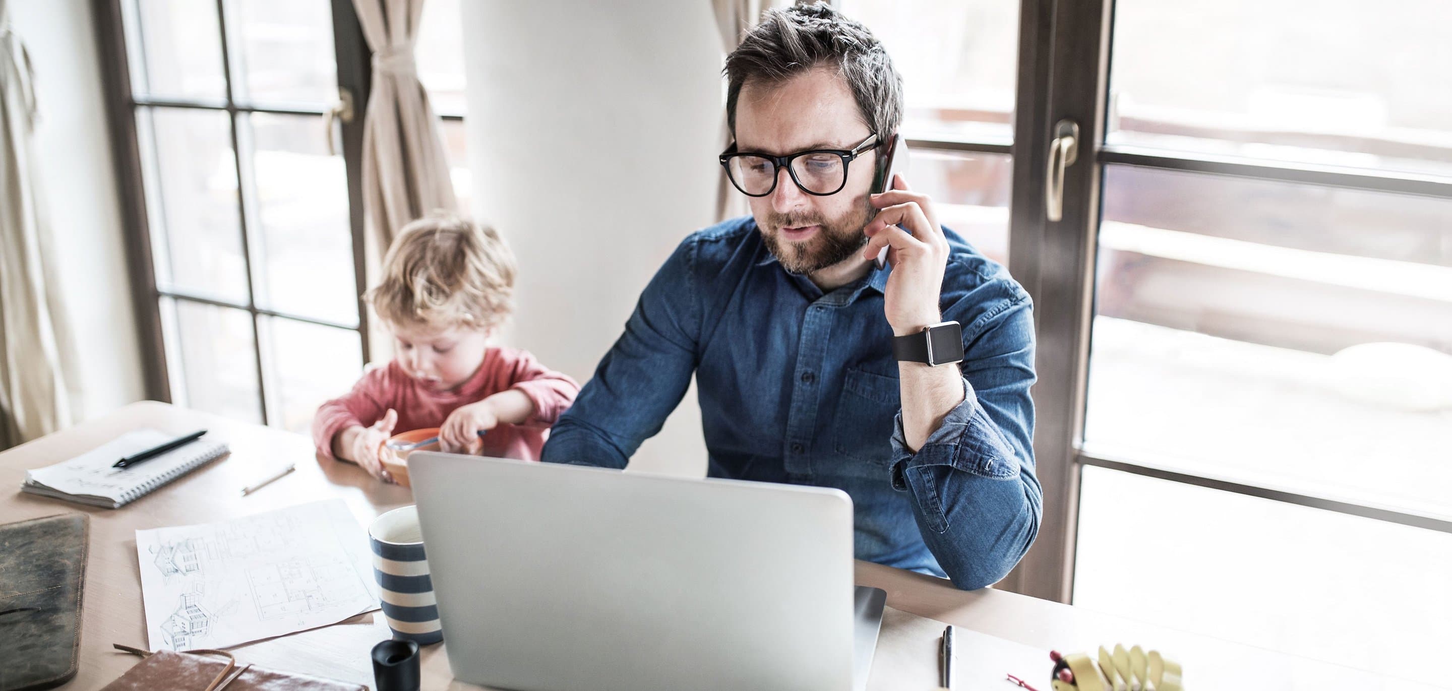 Person arbeitet im Homeoffice an einem Laptop und telefoniert, während ein Kind daneben am Tisch spielt. Auf dem Tisch liegen Notizbuch, Stifte und eine gestreifte Tasse.