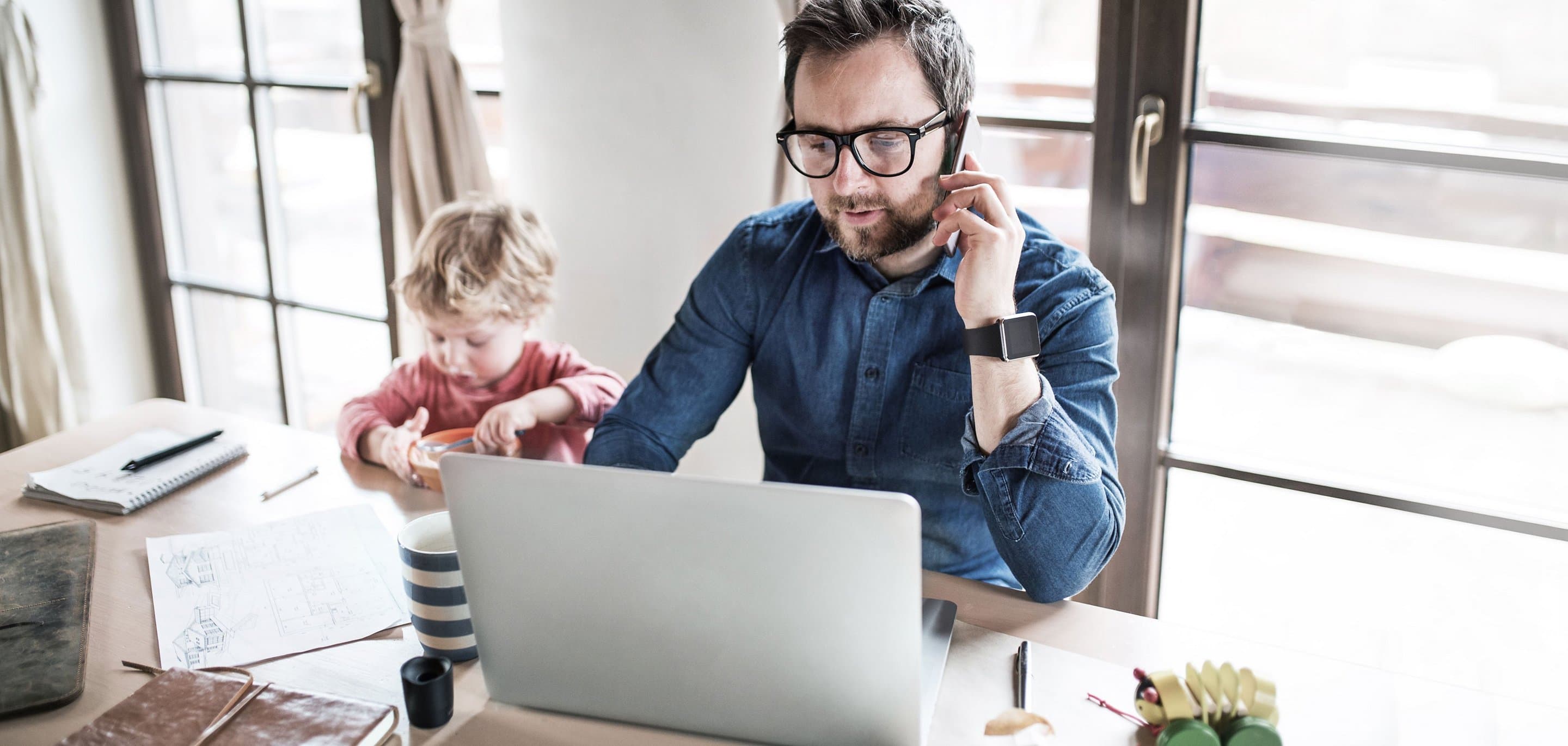 Person arbeitet im Homeoffice an einem Laptop und telefoniert, während ein Kind daneben am Tisch spielt. Auf dem Tisch liegen Notizbuch, Stifte und eine gestreifte Tasse.