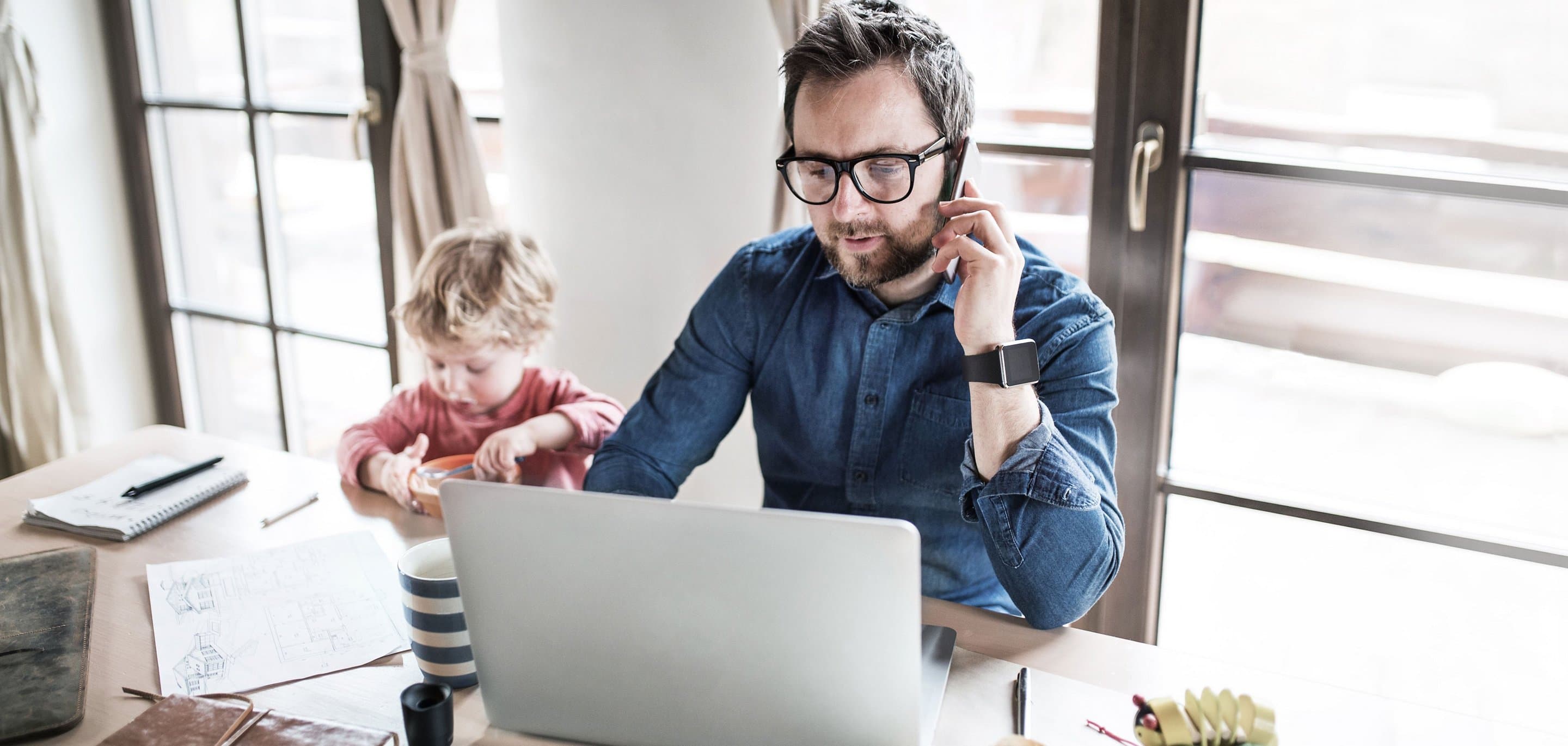 Person arbeitet im Homeoffice an einem Laptop und telefoniert, während ein Kind daneben am Tisch spielt. Auf dem Tisch liegen Notizbuch, Stifte und eine gestreifte Tasse.