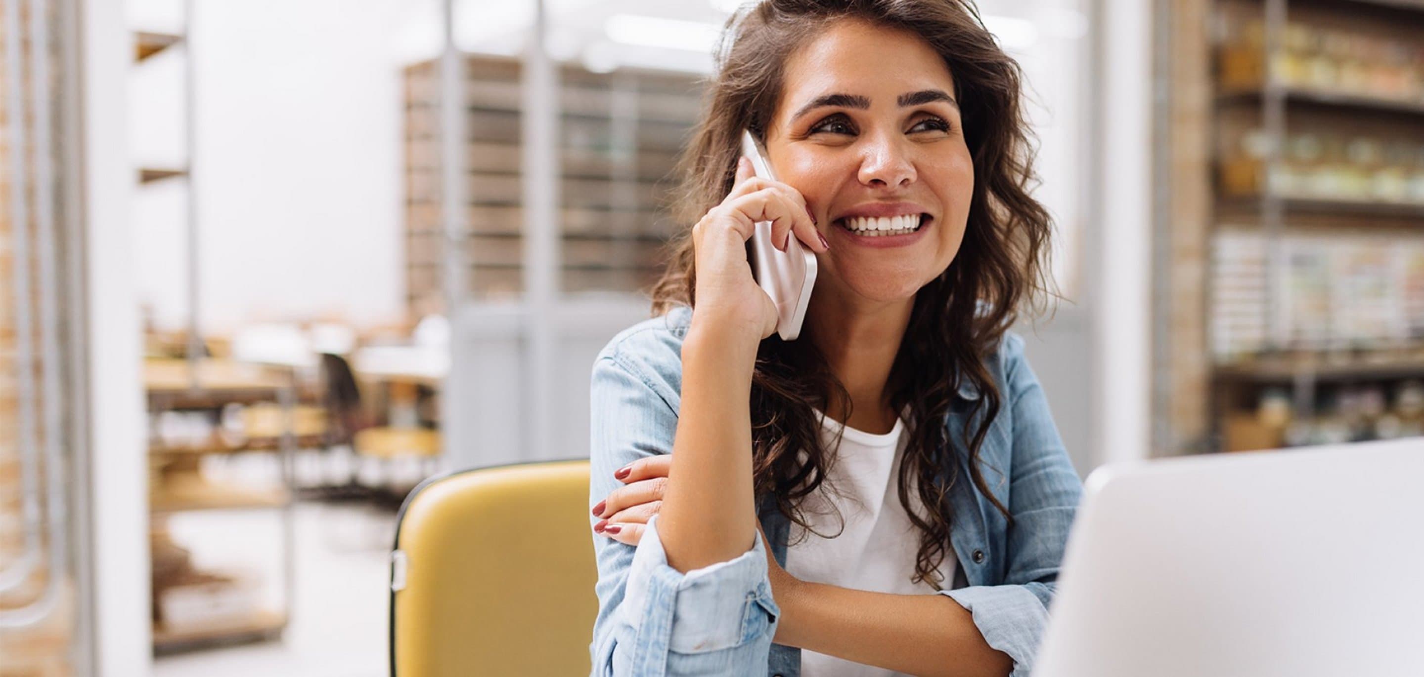 Person sitzt an einem Tisch mit Laptop und führt ein Telefonat, im Hintergrund Regale mit Büchern und Ordnern.