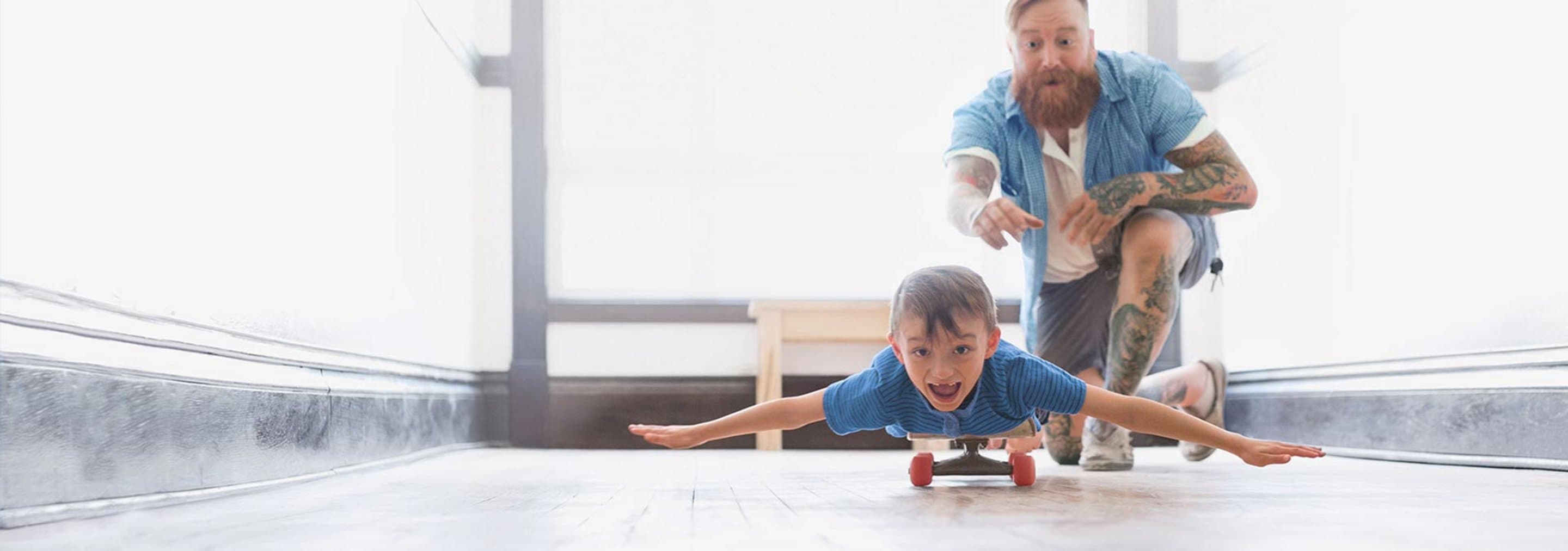 Vater schubst Sohn an, der mit ausgebreiteten Armen auf einem Skateboard liegt.