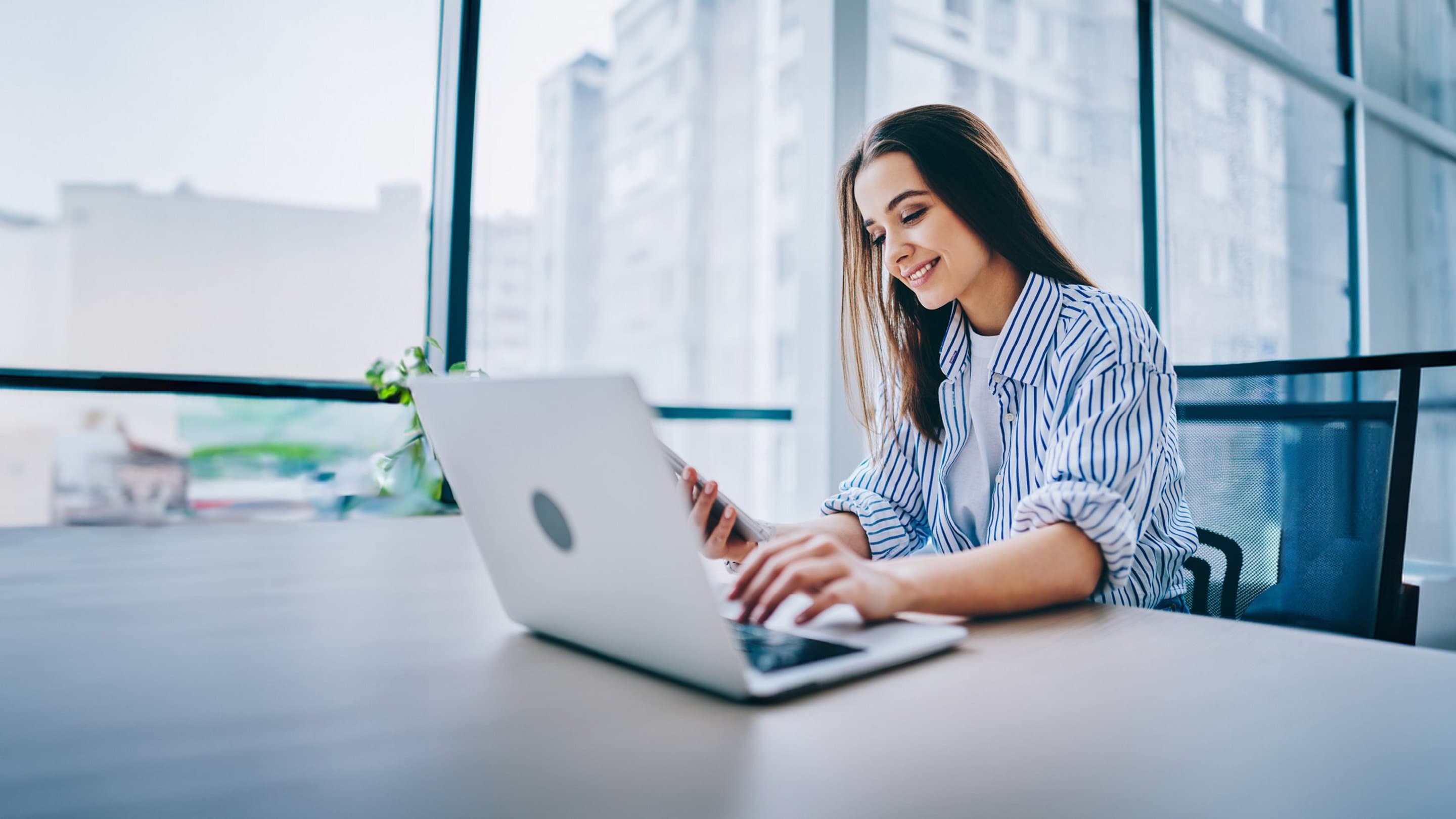 Sichere & werbefreie E-Mail erstellen mit NetMail Frau sitzt am Schreibtisch auf dem ein Laptop steht und schaut auf ihr Handy