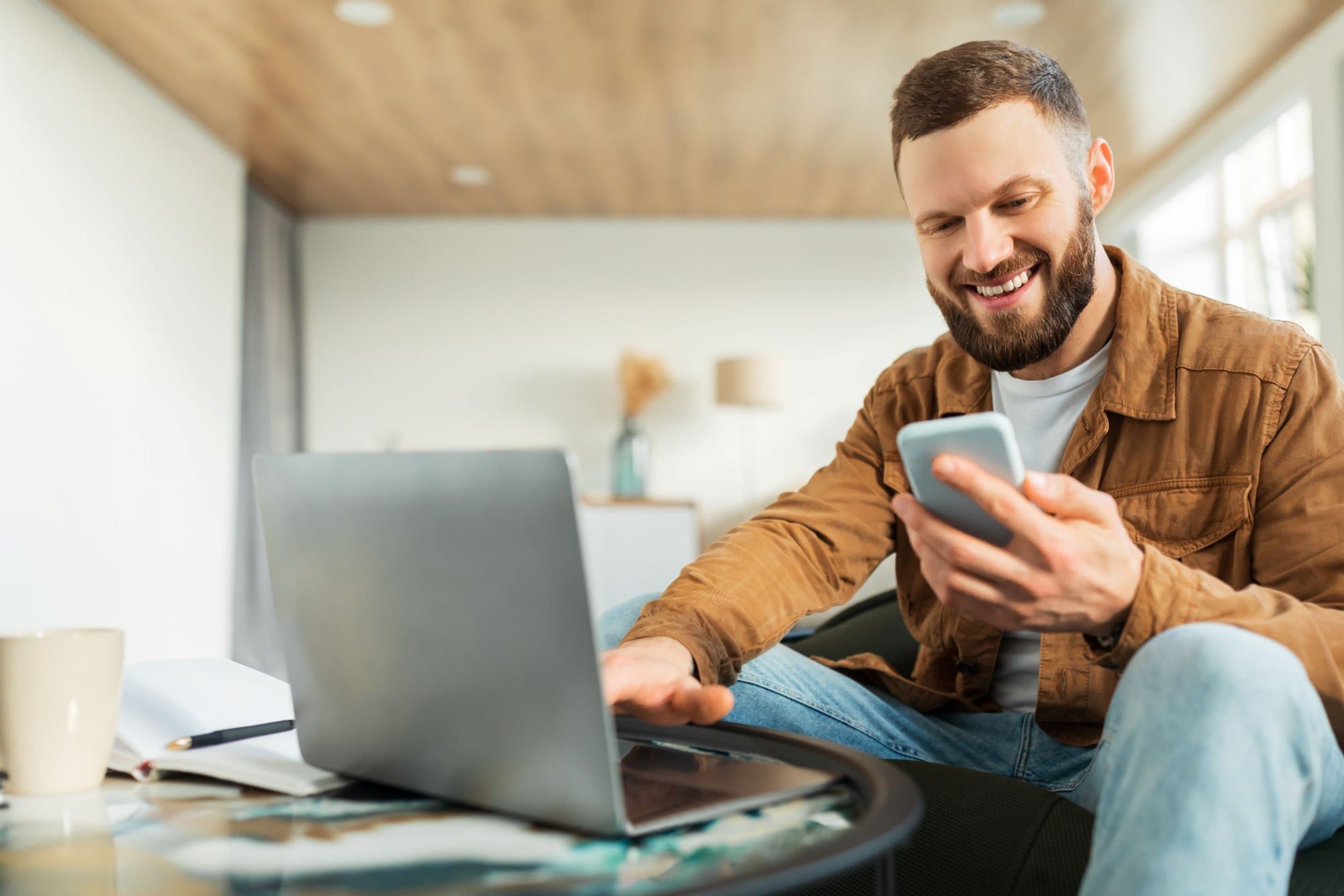 Person sitzt in einem modernen Raum vor einem Laptop und hält ein Smartphone in der Hand, auf dem Tisch steht eine helle Kaffeetasse.