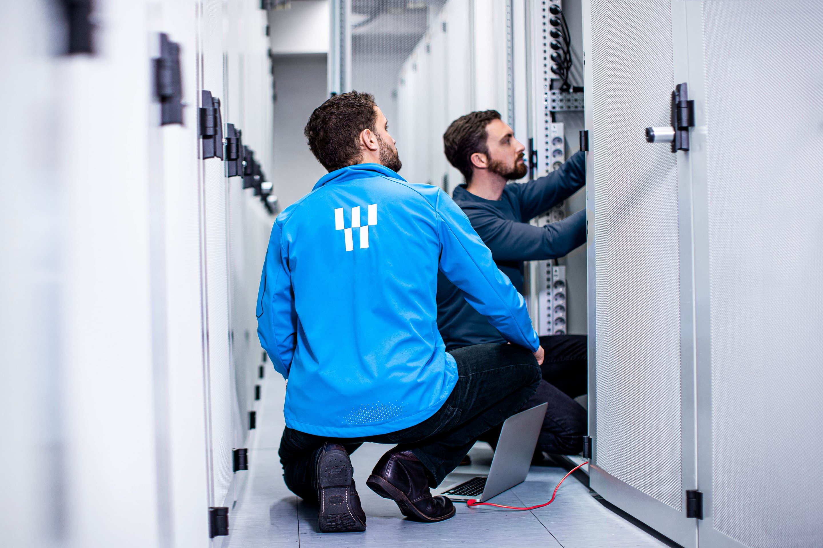 Rechenzentrum von NetAachen Zwei Personen arbeiten in einem Serverraum an offenen Serverschränken, eine trägt eine blaue Jacke mit NetAachen-Logo, auf dem Boden liegt ein Laptop.