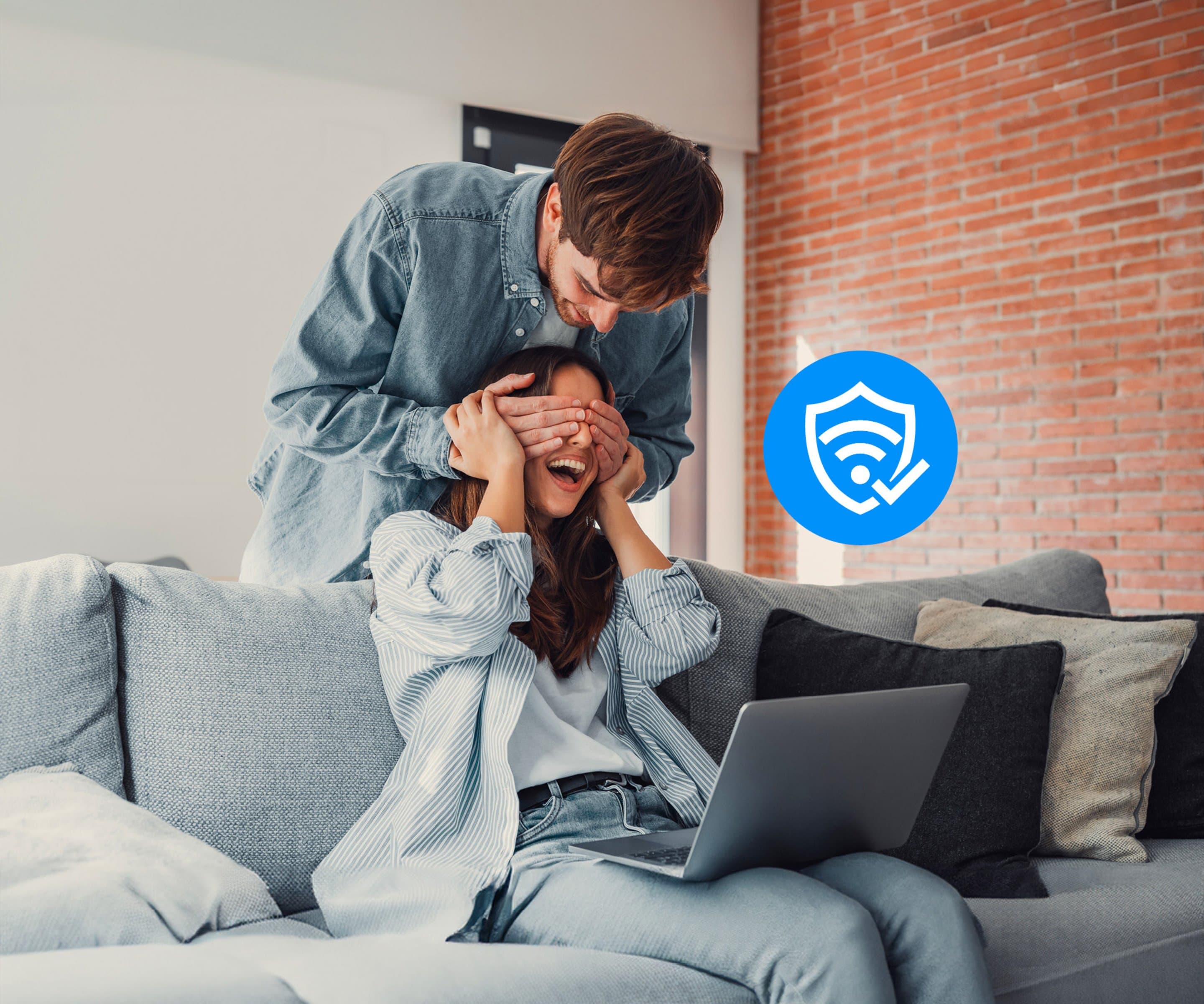 Junge Frau sitzt auf Couch und bekommt Augen von einem jungen Mann zugehalten. Neben dem Paar ist ein Sicherheits-Icon zu sehen.