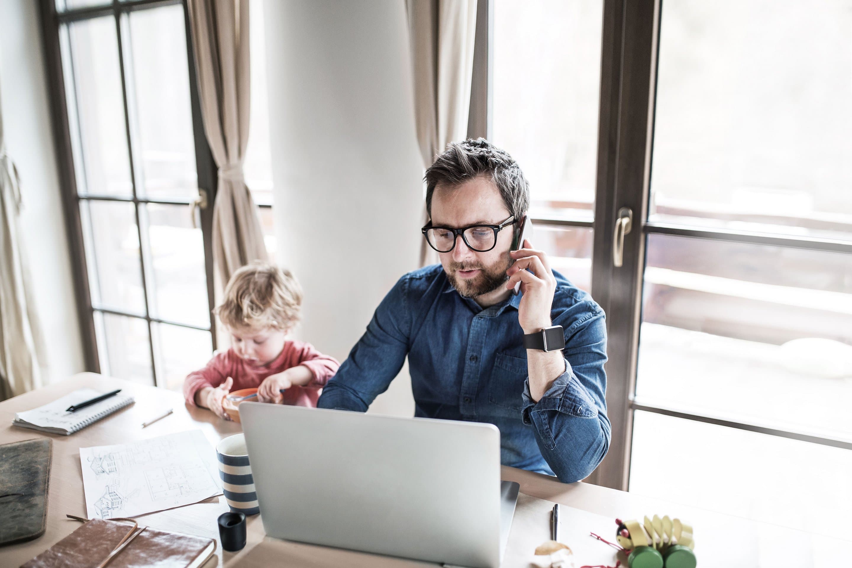 Person arbeitet im Homeoffice an einem Laptop und telefoniert, während ein Kind daneben am Tisch spielt. Auf dem Tisch liegen Notizbuch, Stifte und eine gestreifte Tasse.